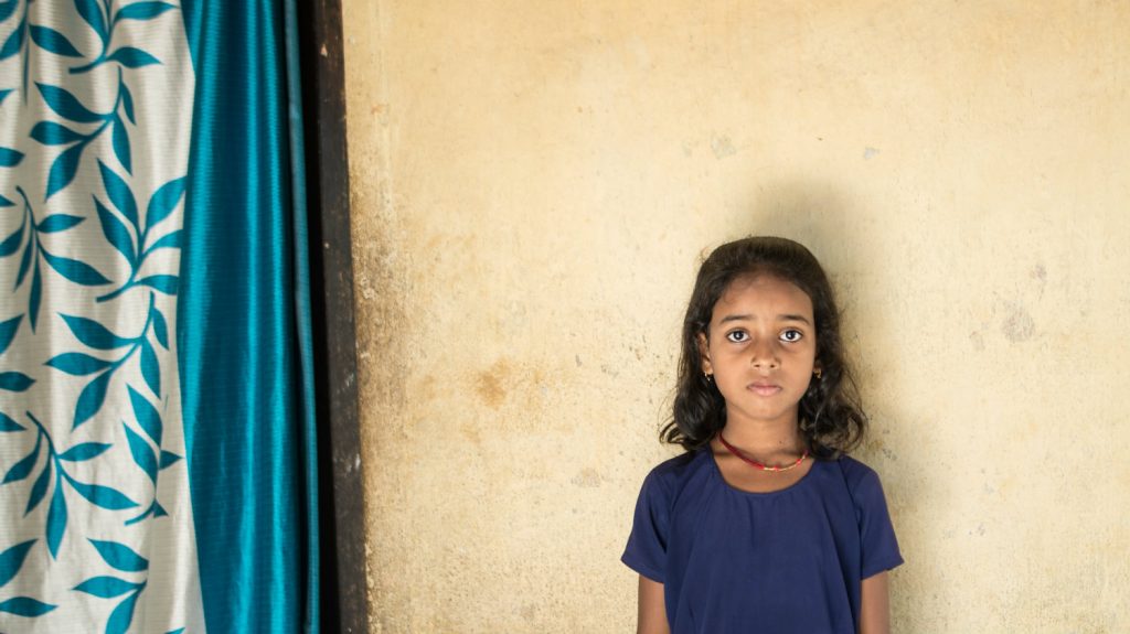 Riya, without glasses, stands against a wall.