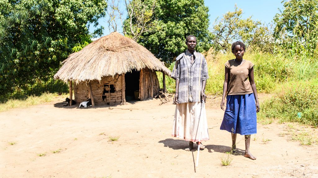 Ayenyo and Lawino stand together outside their hut in Uganda.