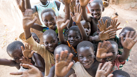 En grupp barn i Ghana ler och vinkar till kameran.