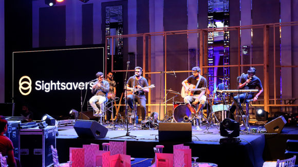A group of musicians play guitar on stage, with Sightsavers banners in the background.
