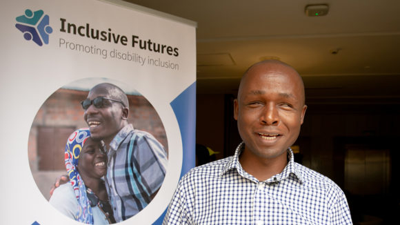 Deus Turyatemba står bredvid en Inclusive Futures-plansch