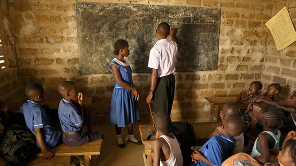 En klar och tydlig bild av ett klassrum i Sierra Leone. Läraren står vid svarta tavlan med en elev medan klasskamraterna tittar på.