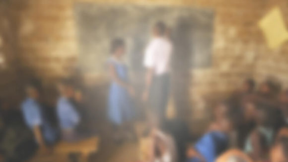 En oskarp bild av ett klassrum i Sierra Leone. Bilden är suddig och otydlig med en vitaktig nyans. Det är svårt att urskilja läraren och svarta tavlan.