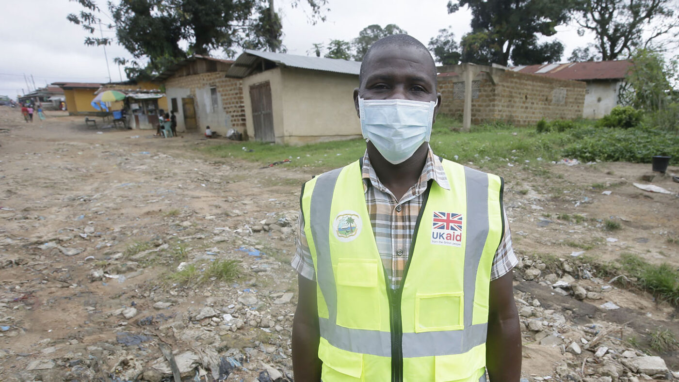 En man med munskydd står på en grusplan med hus i bakgrunden.