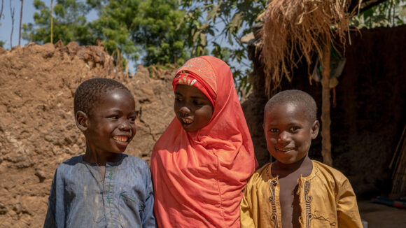 Tre små barn från Nigeria står tillsammans och ler.